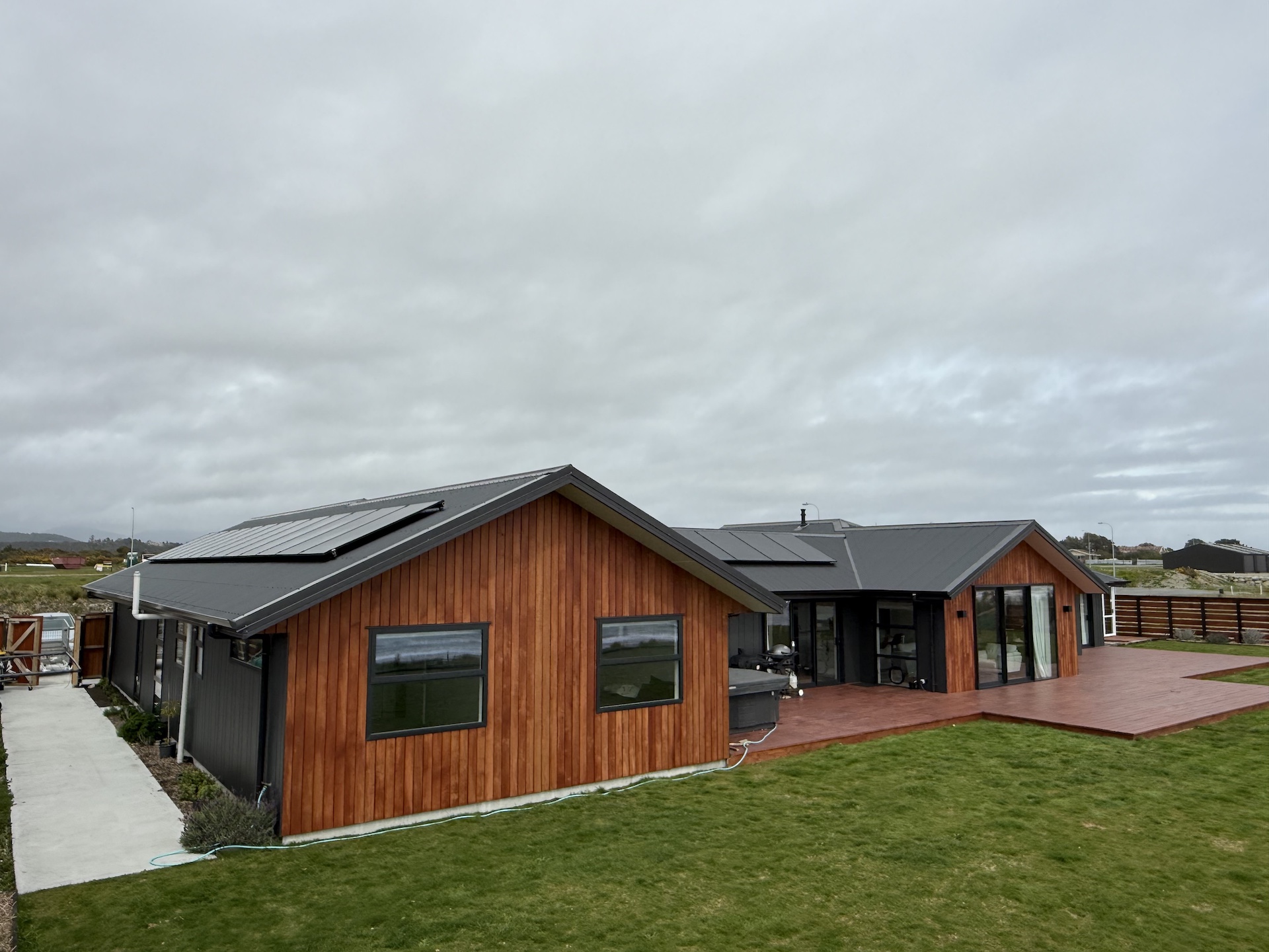 Single-story modern house with wooden siding, large windows, solar panels on the roof, a spacious deck, and a grassy yard under a cloudy sky.