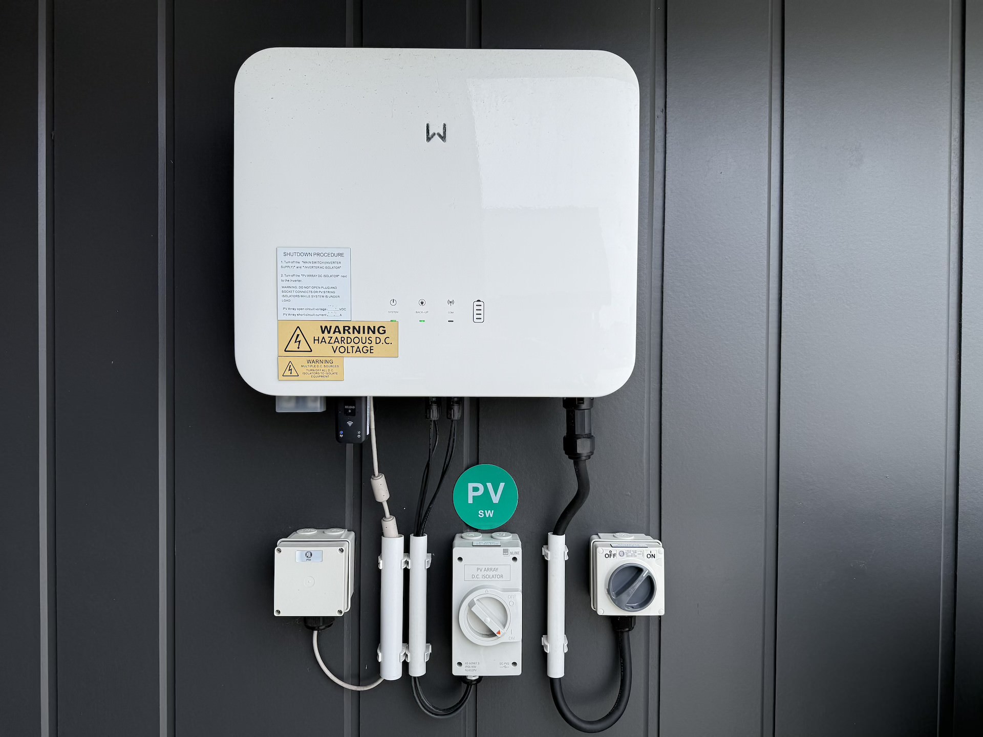 A white solar inverter with warning labels and connected cables mounted on a gray wall, accompanied by control boxes and a green "PV 5kW" label.